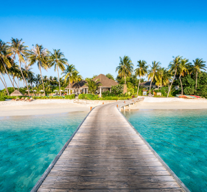 Steg führt über türkisfarbenes Wasser zu einem tropischen Strandresort auf den Malediven, umgeben von Palmen und weißem Sand bei strahlend blauem Himmel.