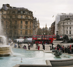 Trafalgar Square in London mit Springbrunnen, Menschenmenge, rotem Doppeldeckerbus und Blick auf Big Ben in der Ferne.
