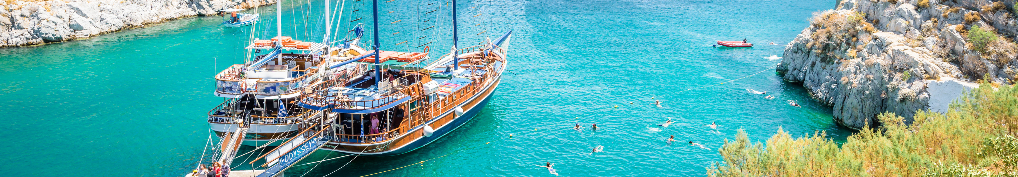 Badebucht mit t&uuml;rkisblauem Wasser, Felsen, Badenden und zwei traditionellen Segelbooten am Steg.