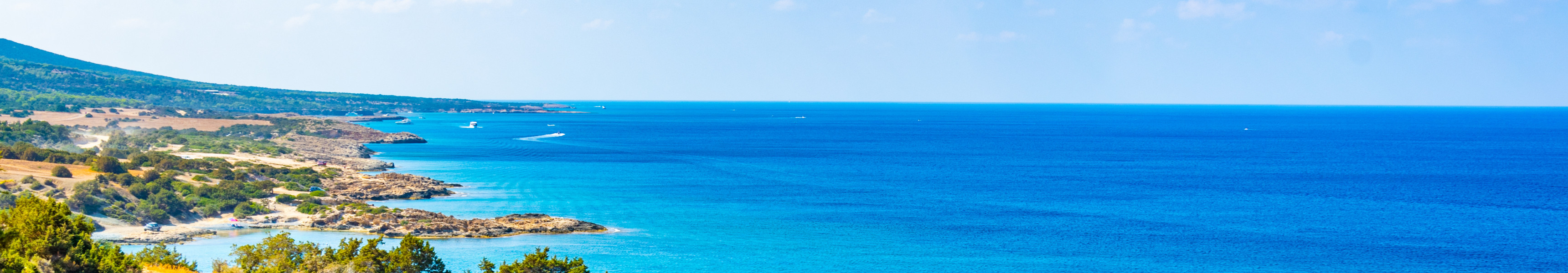 Felsk&uuml;ste mit gr&uuml;n bewachsenen H&uuml;geln und t&uuml;rkisblauem Meer auf Zypern bei klarem Sommerhimmel.