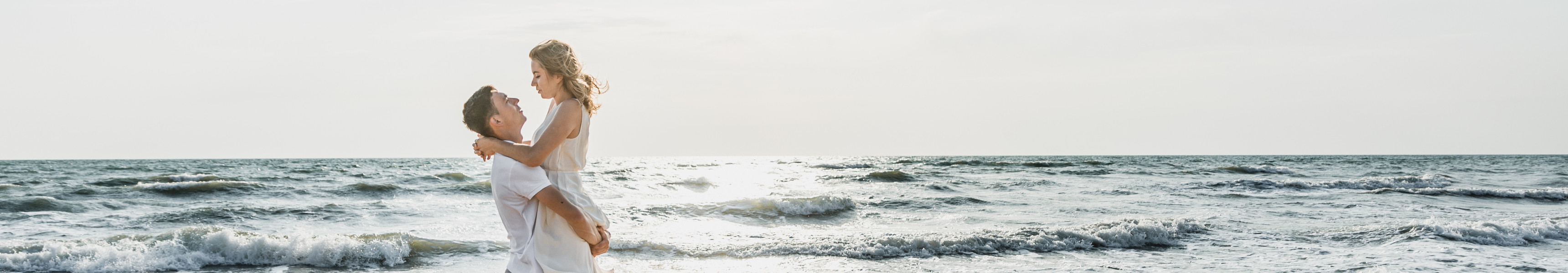 Ein Mann hebt eine Frau in wei&szlig;em Kleid am Strand hoch, w&auml;hrend beide lachend das Meer im Hintergrund und die untergehende Sonne genie&szlig;en.