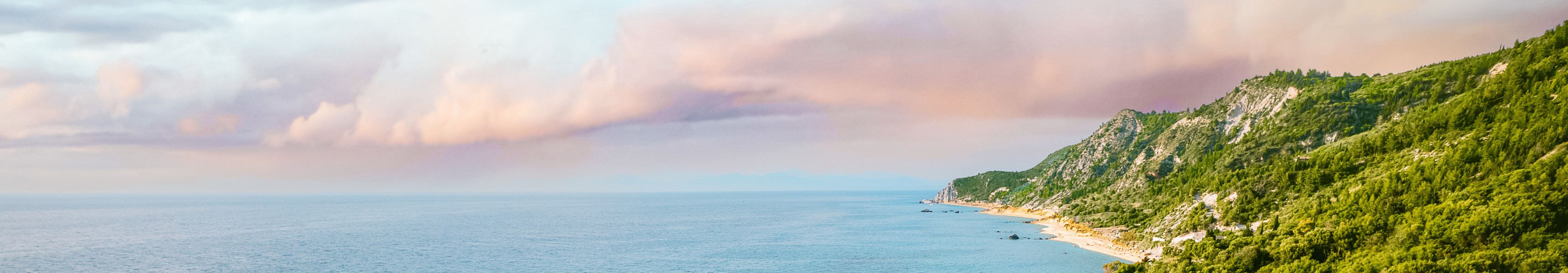 Blick auf das türkisfarbene Meer in Griechenland mit einem Sandstrand, großen Felsen, grünen Hügeln im Hintergrund und Reihen von blauen Sonnenliegen am Ufer.