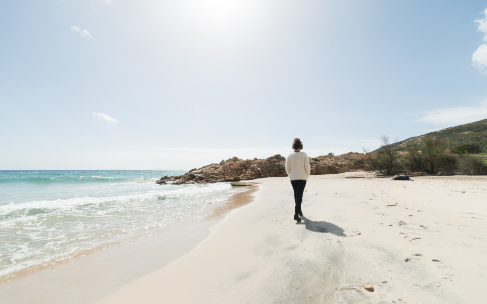 Eine Person spaziert am ruhigen Sandstrand entlang, w&auml;hrend die Sonne scheint &ndash; eine Methode zur Anpassung des Tagesablaufs zur Vermeidung von Jetlag.