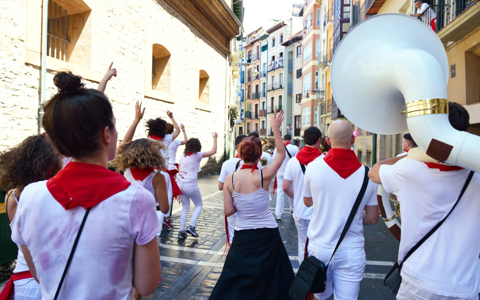 Menschen in wei&szlig;er Kleidung mit roten Halst&uuml;chern tanzen und feiern auf einer engen Stra&szlig;e zwischen historischen Geb&auml;uden, begleitet von einer Blaskapelle.