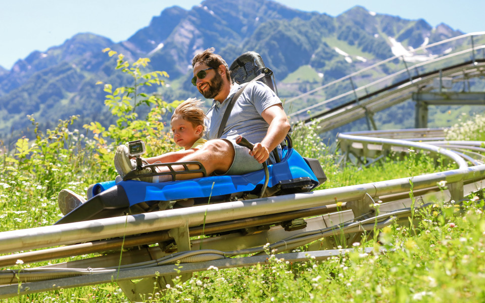 Vater und Kind fahren bei Sonnenschein in einem blauen Sommerrodel auf einer Schienenbahn durch gr&uuml;ne Almwiesen, im Hintergrund hohe Berge mit Schneeresten.